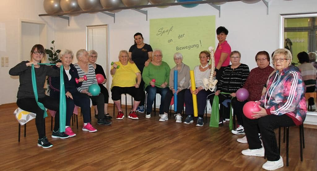 Eine Kursgruppe bei Rolling Pritschen: Frauen sitzen mit Bändern, Bällen und Hanteln nebeneinander und schauen freundlich in die Kamera